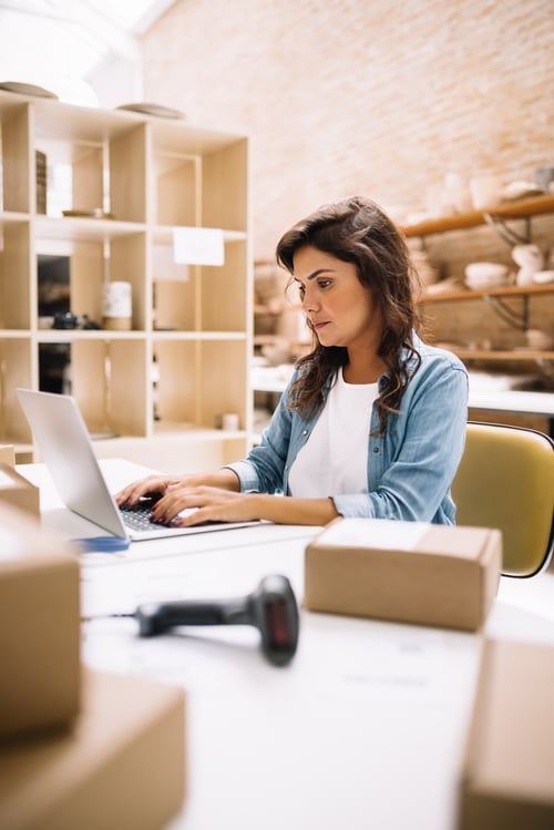 Woman working on laptop in a creative workspace, surrounded by packaging boxes, emphasizing digital marketing and brand management.